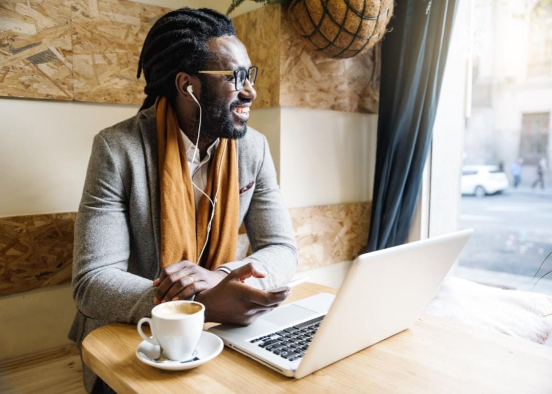 A smiling person at a coffee shop with headphones and a computer.