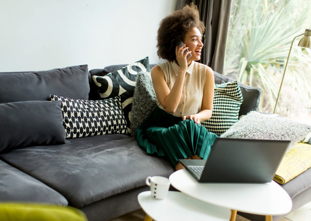 A person in a green silk skirt and beige top sitting on the couch talking on the phone in front of her laptop.